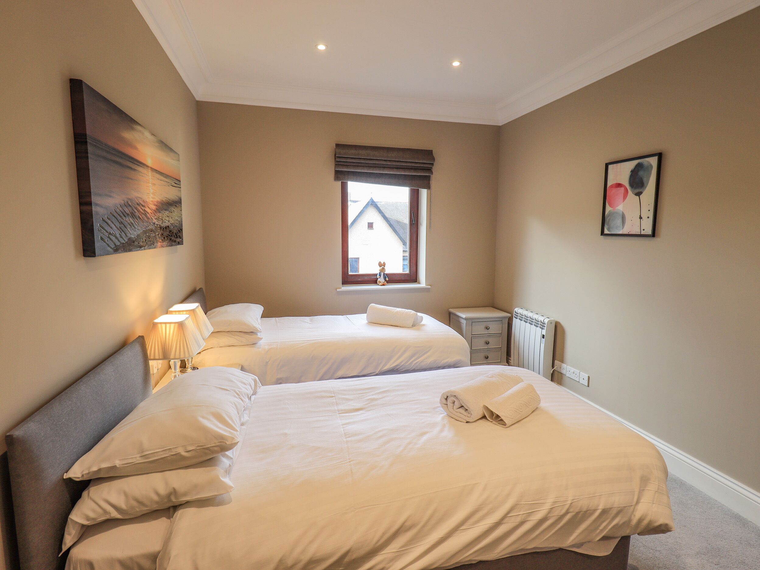 A bedroom with two beds, a nightstand, a radiator, and a window with a view of a house.
