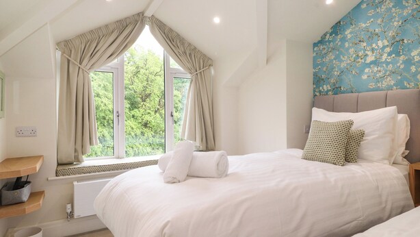 A bedroom with a large bed, a window with curtains, a radiator, a shelf, and a wall with a floral pattern. - Jenever Cottage (Ambleside)