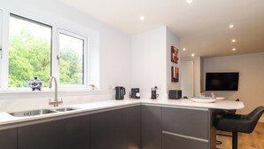 A modern kitchen with a large window, a sink, a coffee maker, and a bar area with a TV. - Jenever Cottage (Ambleside)