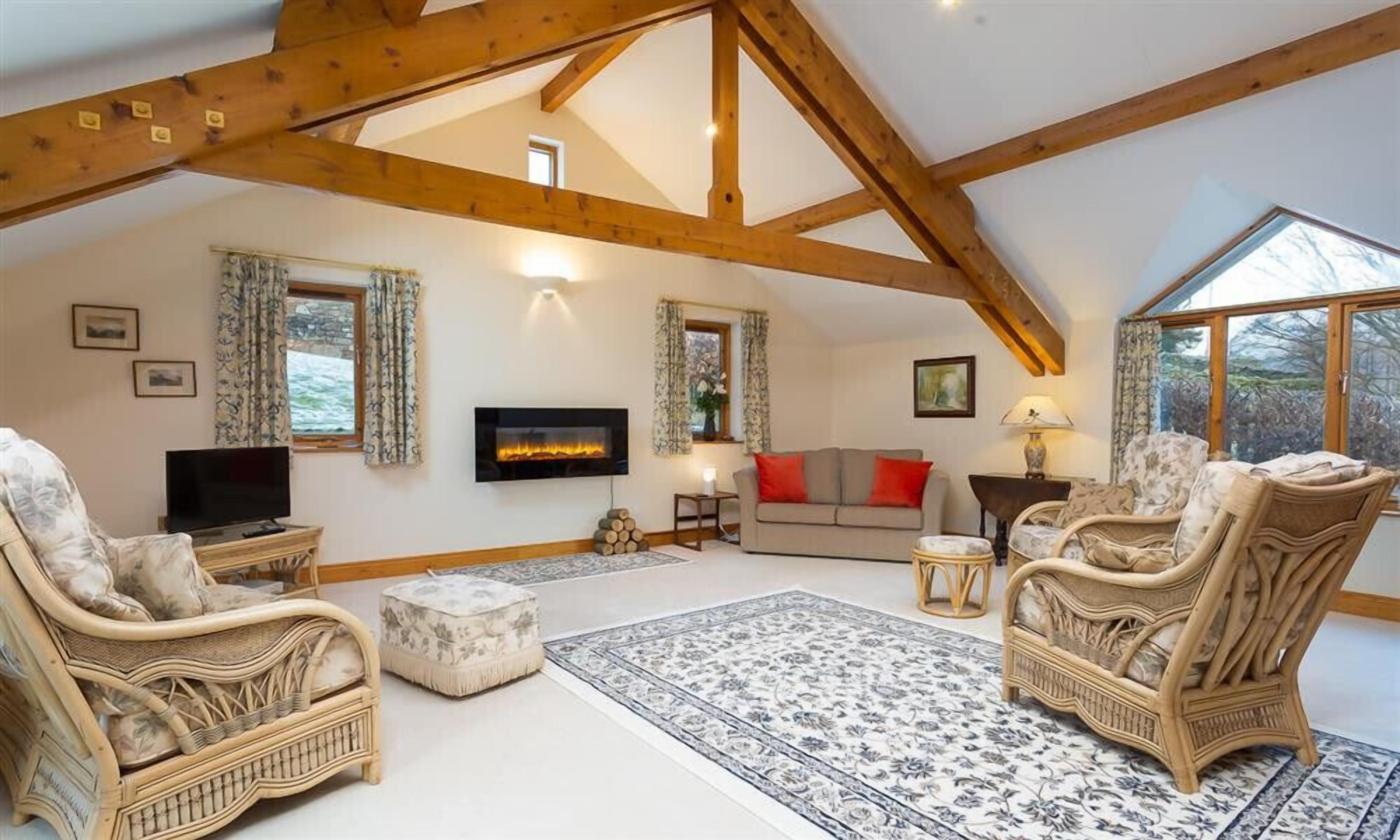A spacious living room with high ceilings and exposed wooden beams, a fireplace, comfortable seating, and large windows offering natural light.