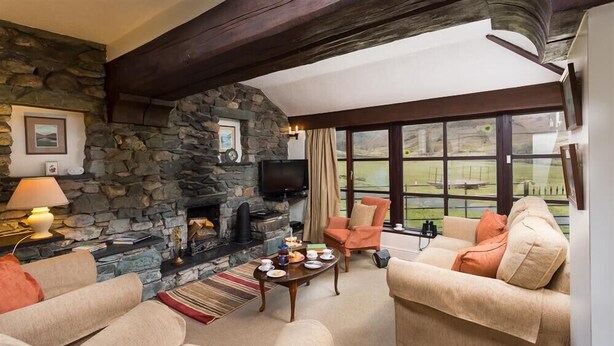A cozy living room with a stone fireplace, comfortable seating, and a large window offering a view of the outdoors. - Thirlmere Cottage (Keswick)