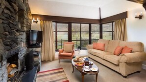 A cozy living room with a stone fireplace, a television, a sofa, and a large window offering a view of the outdoors. - Thirlmere Cottage (Keswick)