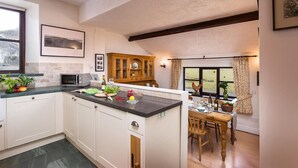 A kitchen with white cabinets, a wooden dining table, and a view of the outdoors through a window. - Thirlmere Cottage (Keswick)