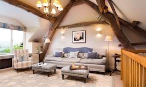 A cozy loft bedroom with exposed wooden beams, a grey sofa, two armchairs, and a wooden railing.
