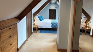 A bedroom with a sloped ceiling, wooden beams, a bed with blue bedding, a bedside table with a lamp, and a wooden dresser. - Dancing Beck Cottage (Keswick)