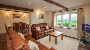 A living room with a brown leather sofa, a wooden coffee table, and a dining area with a table and chairs. - Dancing Beck Cottage (Keswick)
