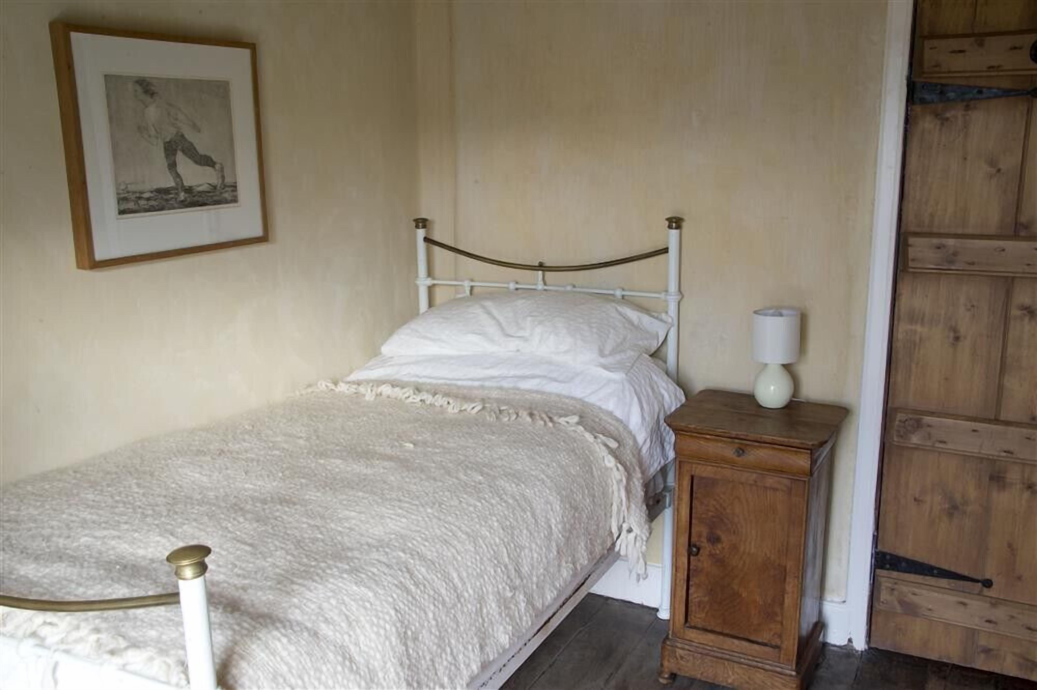 A single bed with a white mattress and a white blanket, a wooden nightstand with a lamp, and a framed picture on the wall.