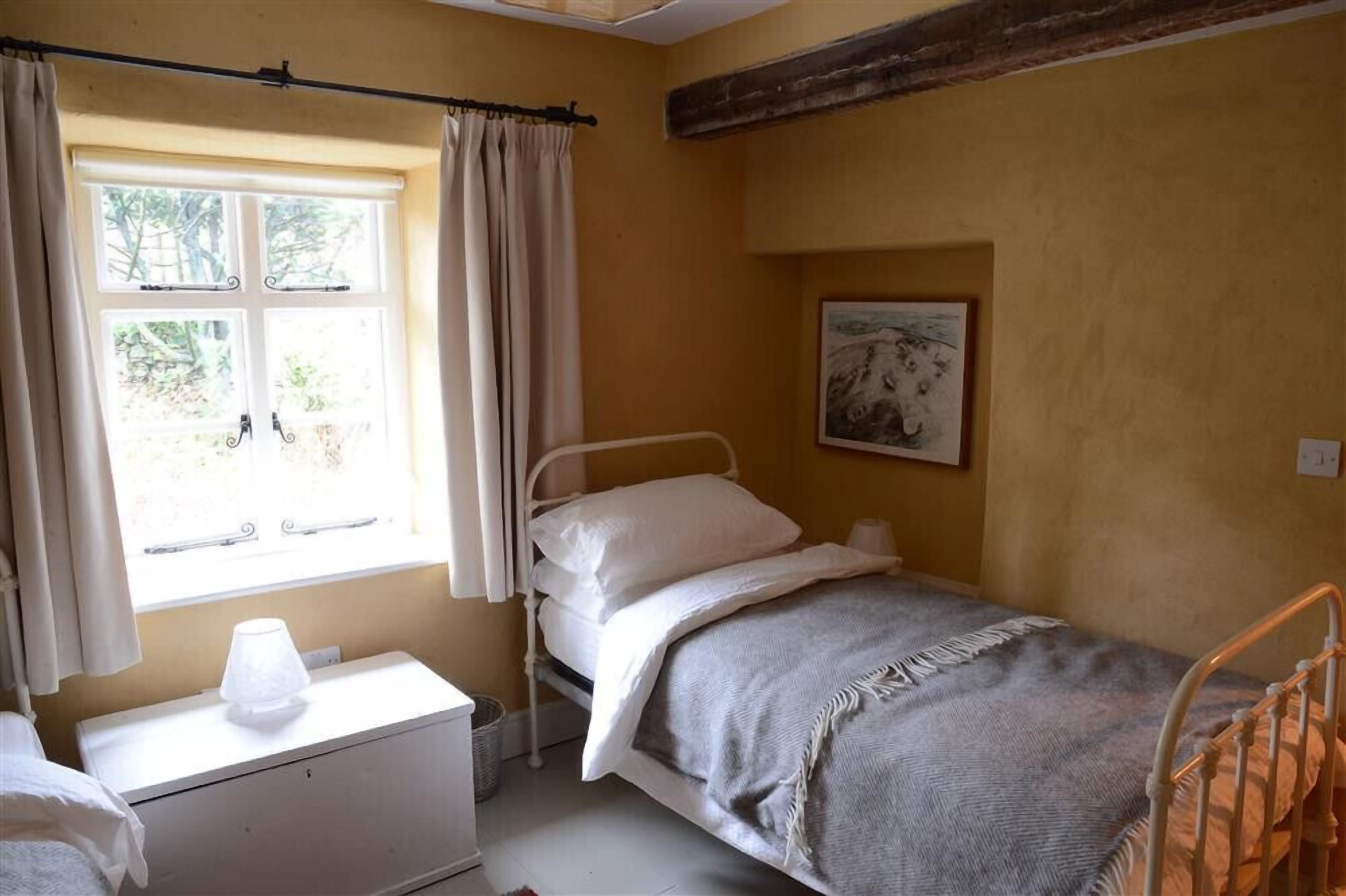 A single bed with a headboard, a bedside table with a lamp, a window with curtains, and a framed picture on the wall.