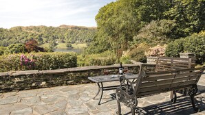 A stone patio with a metal table and bench overlooking a scenic valley with a lake and hills. - Wetherlam (Ambleside)