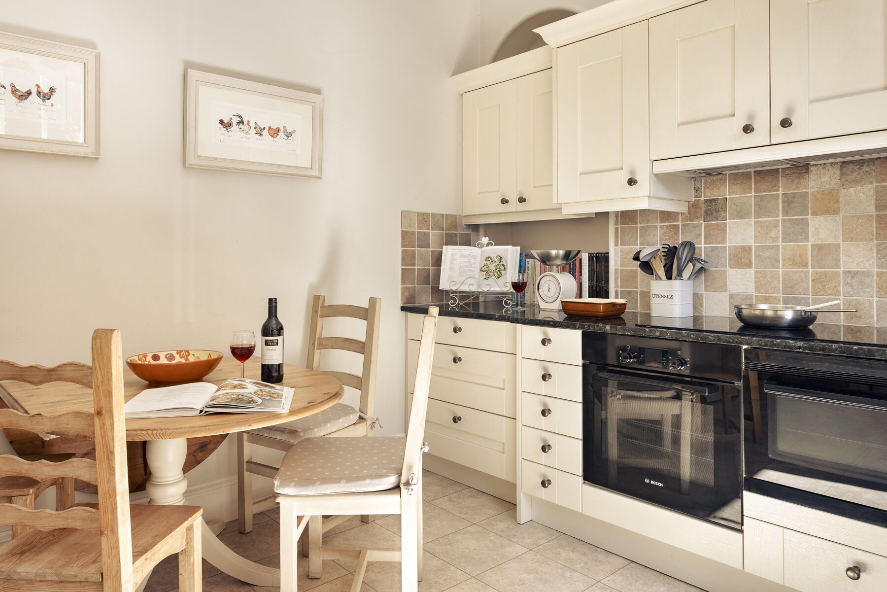 Una cocina con armarios blancos, un horno Bosch, una mesa de comedor con sillas y cuadros en la pared.