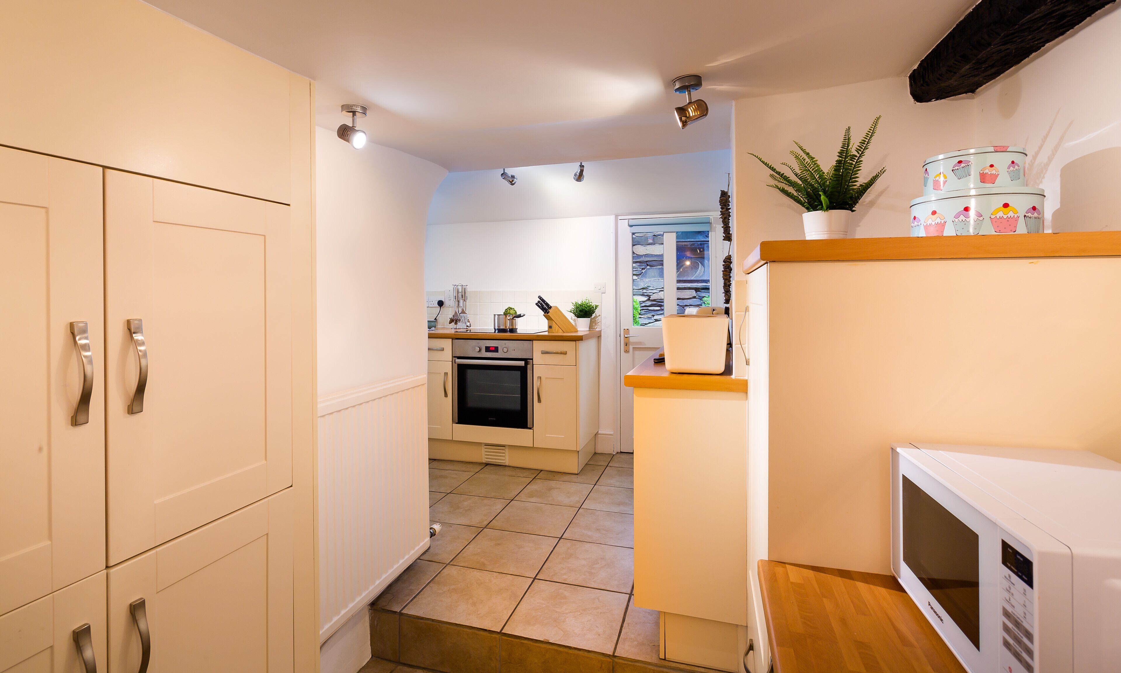 A narrow hallway leading to a kitchen with a built-in oven, a microwave, and a small countertop with a plant and decorative items.