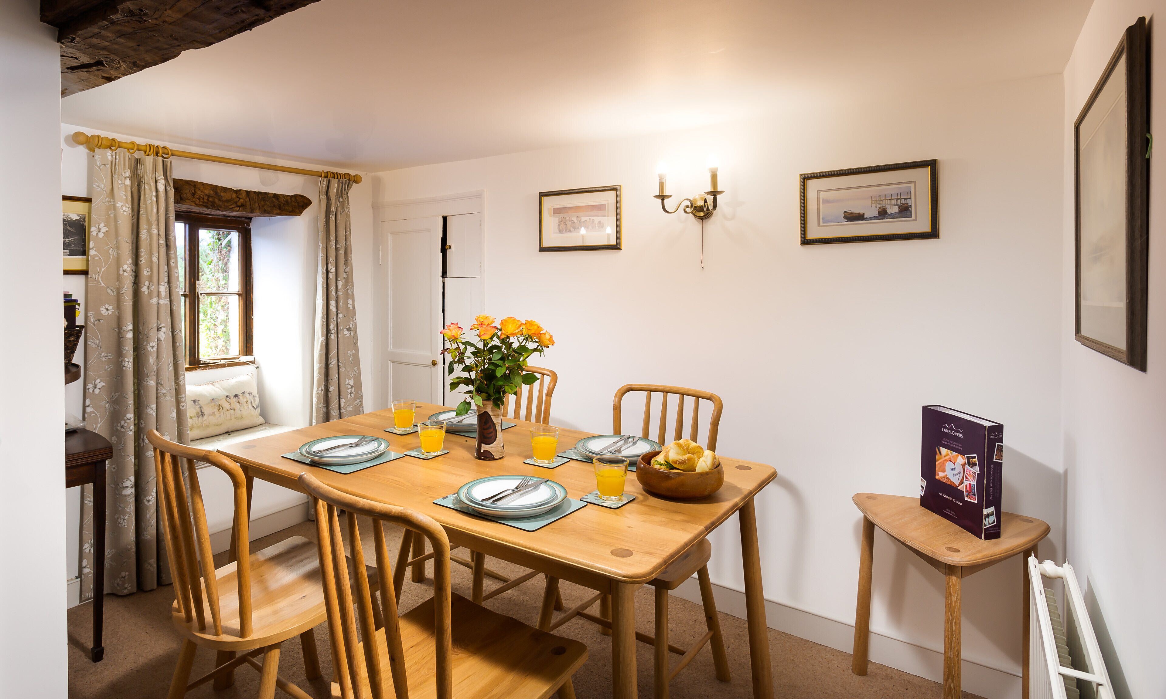 A dining area with a wooden table set for four, chairs, a vase with flowers, and a box of Layloaves bread.