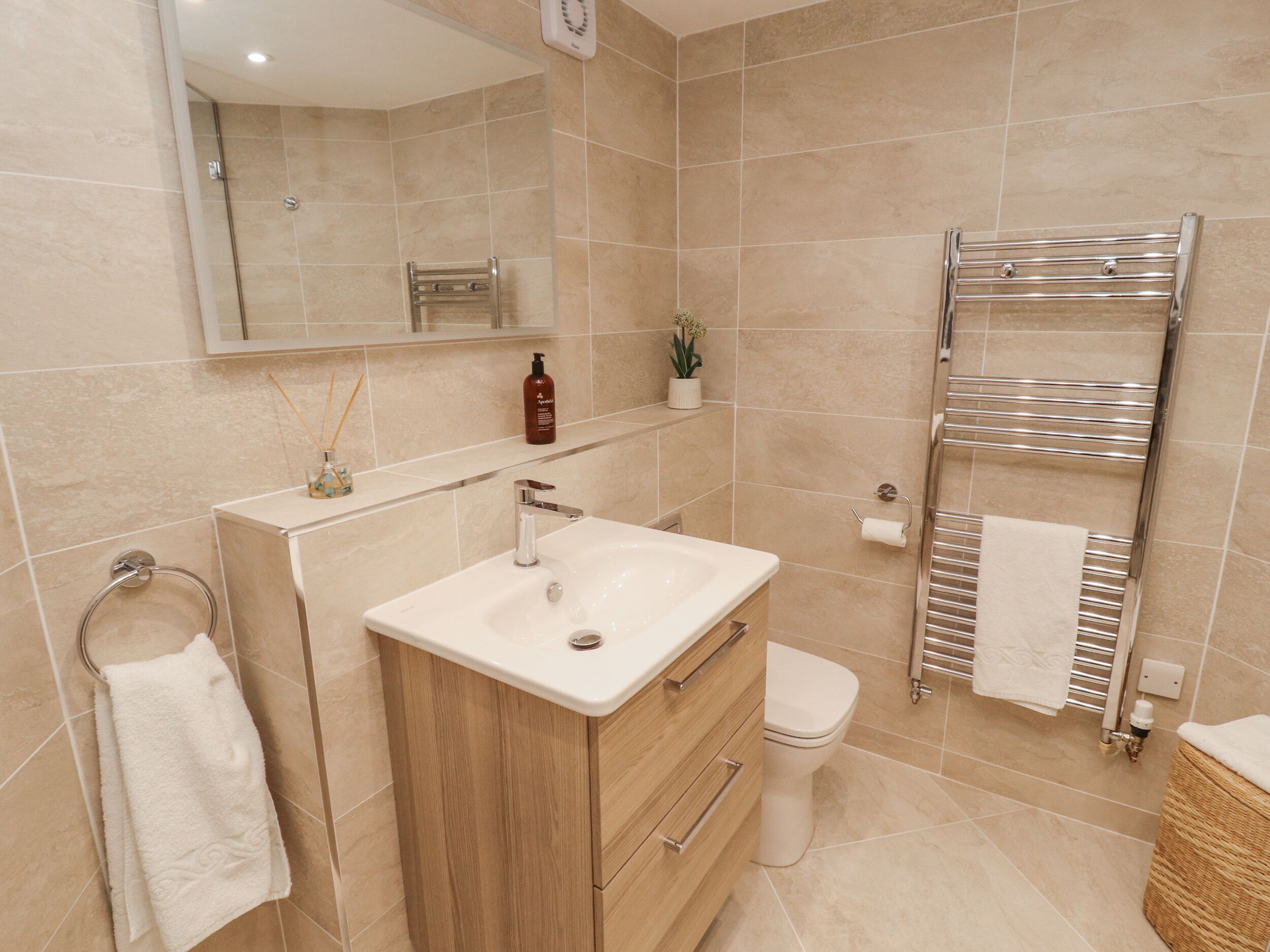 A modern bathroom with a beige tiled wall, a white sink, a wooden vanity, a toilet, and a towel rack.