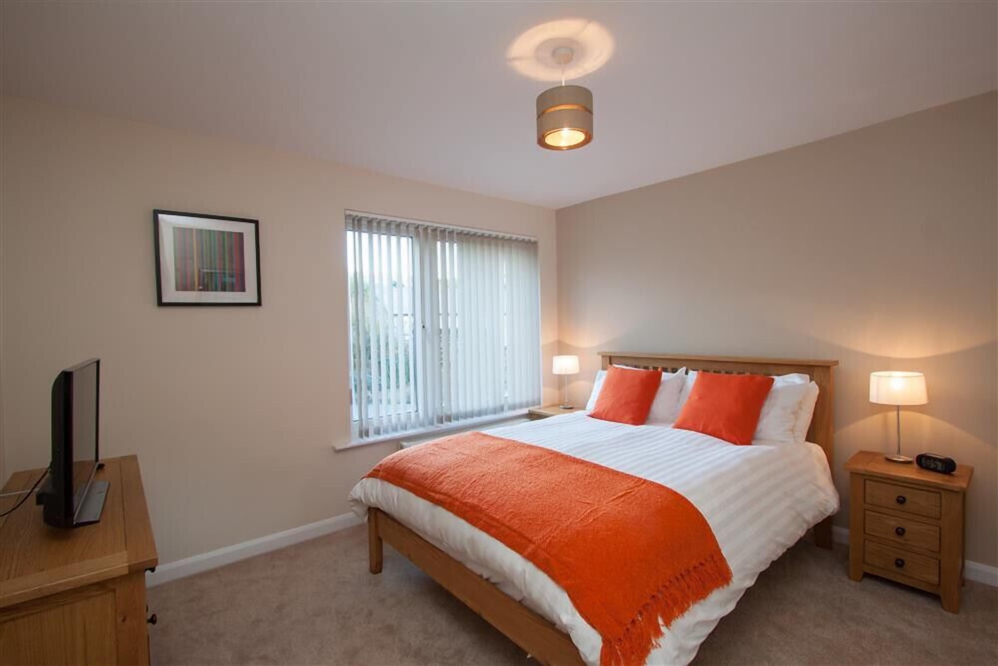 A bedroom with a bed, bedside tables, a TV, a window with blinds, and a ceiling light.