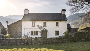 A two-story white house with a stone wall in front, a green door, and a stone chimney. - Yew Tree Farm (Coniston)