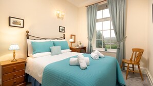 Un lit bien fait, recouvert d’une couvre-lit turquoise et agrémenté d’oreillers, une table de chevet en bois avec une lampe, une chaise en bois et une fenêtre avec des rideaux.