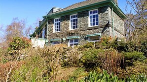 Une maison en pierre dotée de nombreuses fenêtres et d’un toit végétalisé, entourée d’une végétation dense.