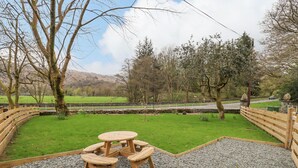 A picnic area with a round table and benches, surrounded by a gravel path and wooden fence, overlooking a green field and a road. - North Lodge (Ambleside)