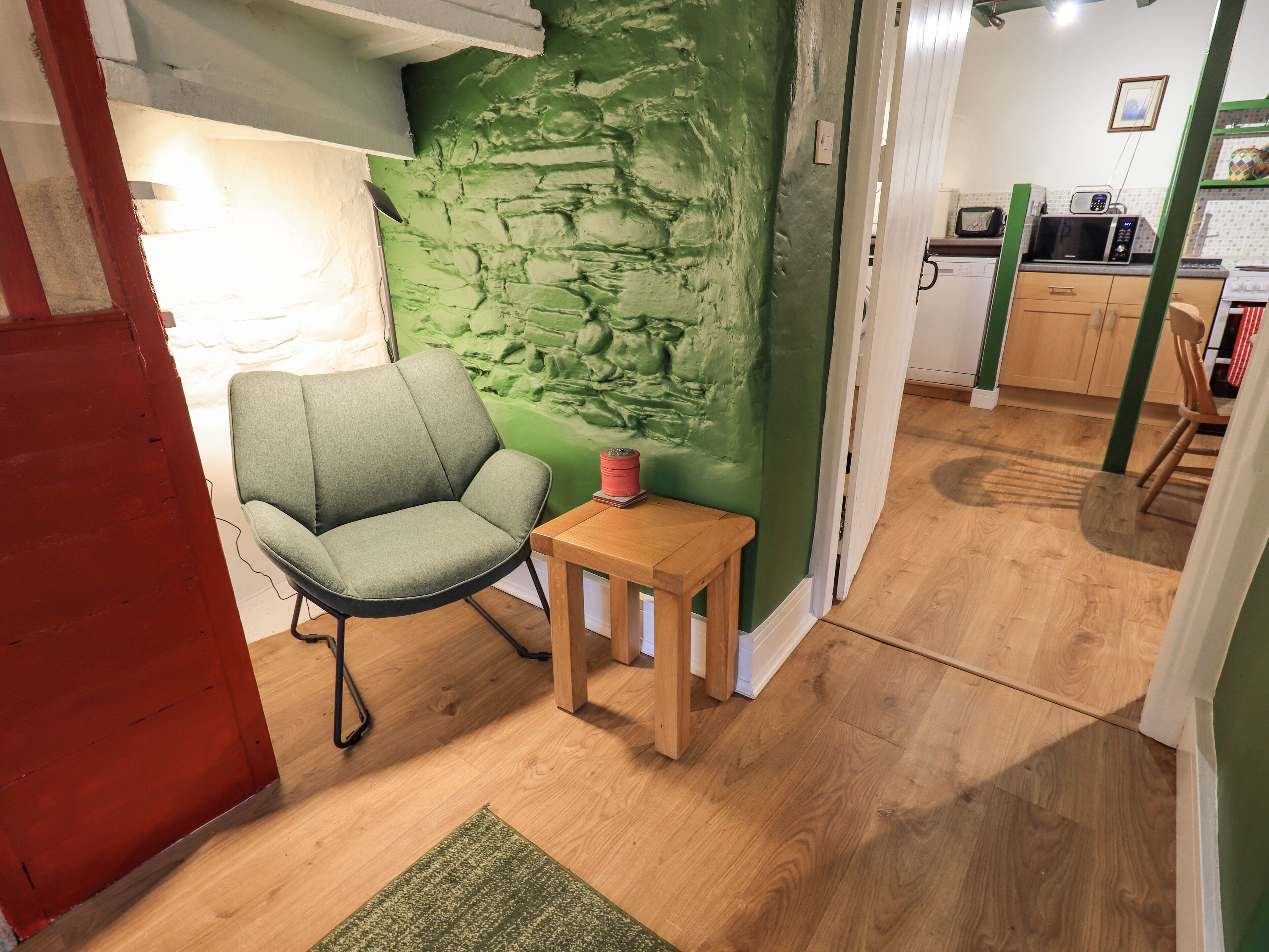 A room with a green textured wall, a grey armchair, a small wooden table, and a kitchen area in the background.