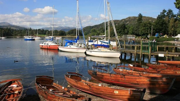 A marina with various boats docked, including sailboats and rowboats, with a calm lake and hills in the background. - Over Ghyll (Ambleside)