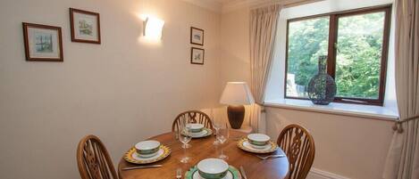 A dining room with a round wooden table set for four, surrounded by wooden chairs. There are framed pictures on the wall, a lamp on the table, and a window with curtains.