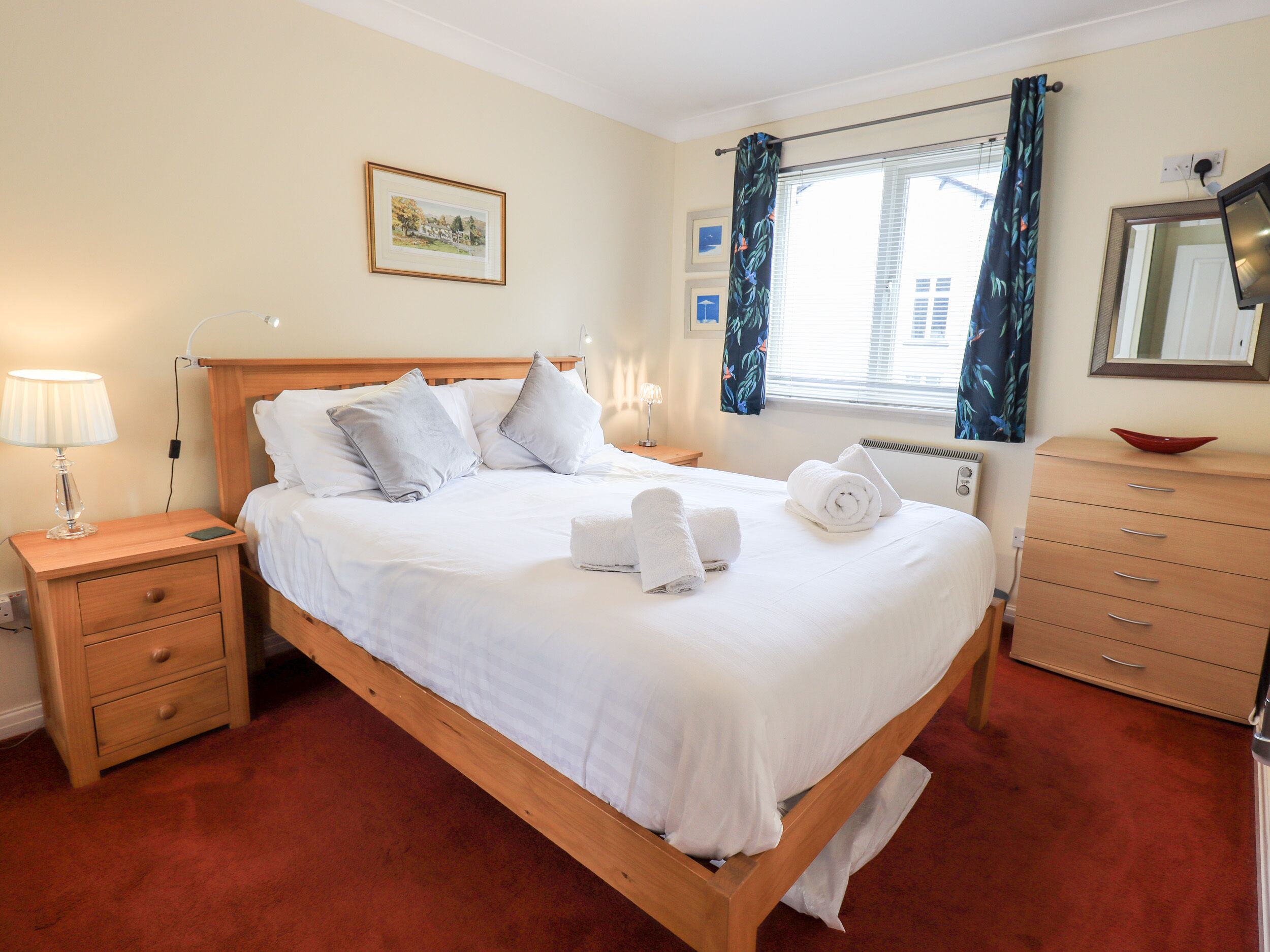 A neatly made bed with white linens, a wooden headboard, and two pillows. There is a nightstand with a lamp and a drawer, a dresser with a mirror, and a window with curtains.