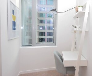 A room with a desk, chair, white shelving unit, and a window with a view of a building.