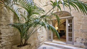 A stone-walled courtyard with a large potted plant, a tiled floor, and a doorway leading to an interior space with furniture. - Rose Of Rhodes Palazzo  (Valletta)