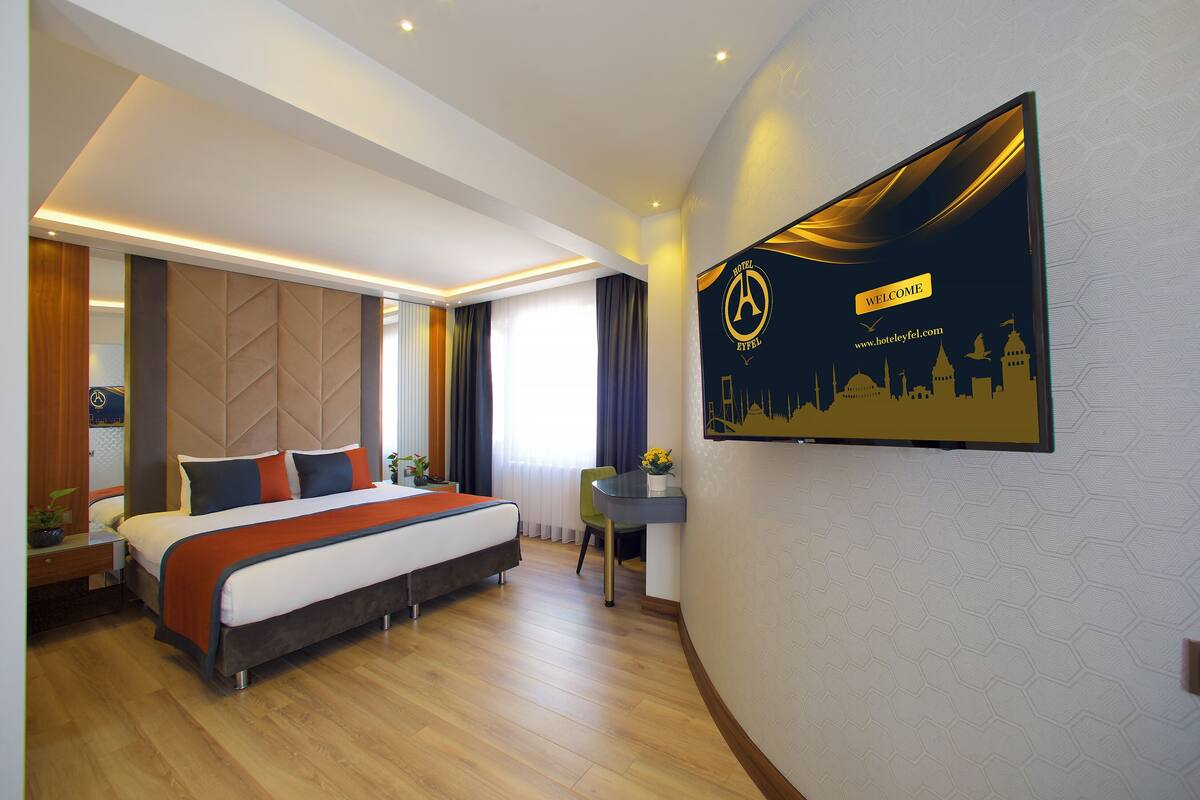 A modern hotel room with a large bed, a desk, and a wall-mounted TV displaying the hotel's logo and website.