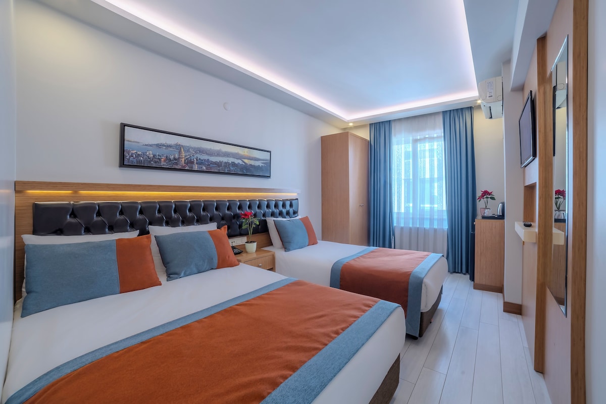 A hotel room with two beds, a cityscape painting, a large window with curtains, and a wooden wardrobe.