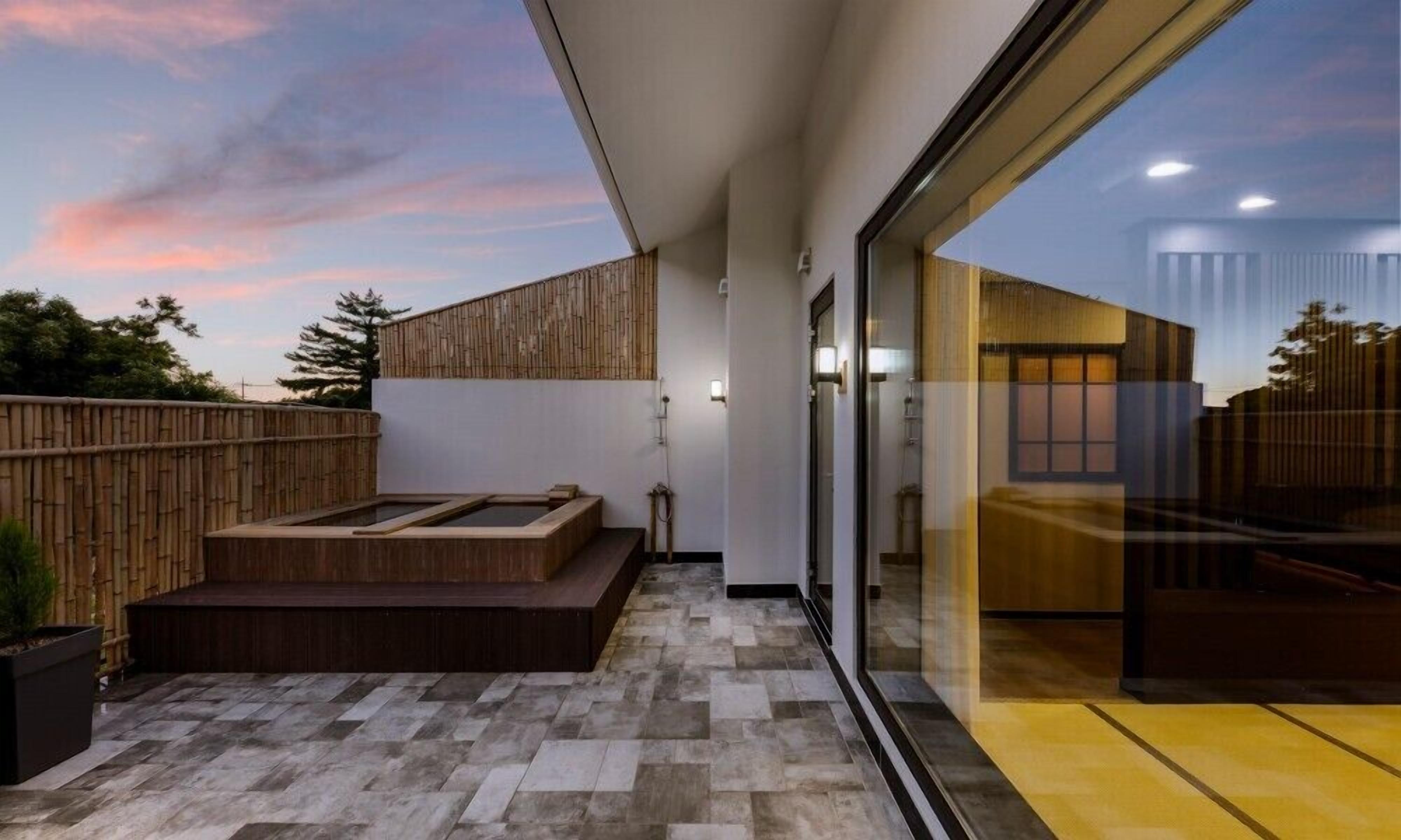 A modern outdoor patio with a wooden deck, a hot tub, and a glass wall leading to an interior space.