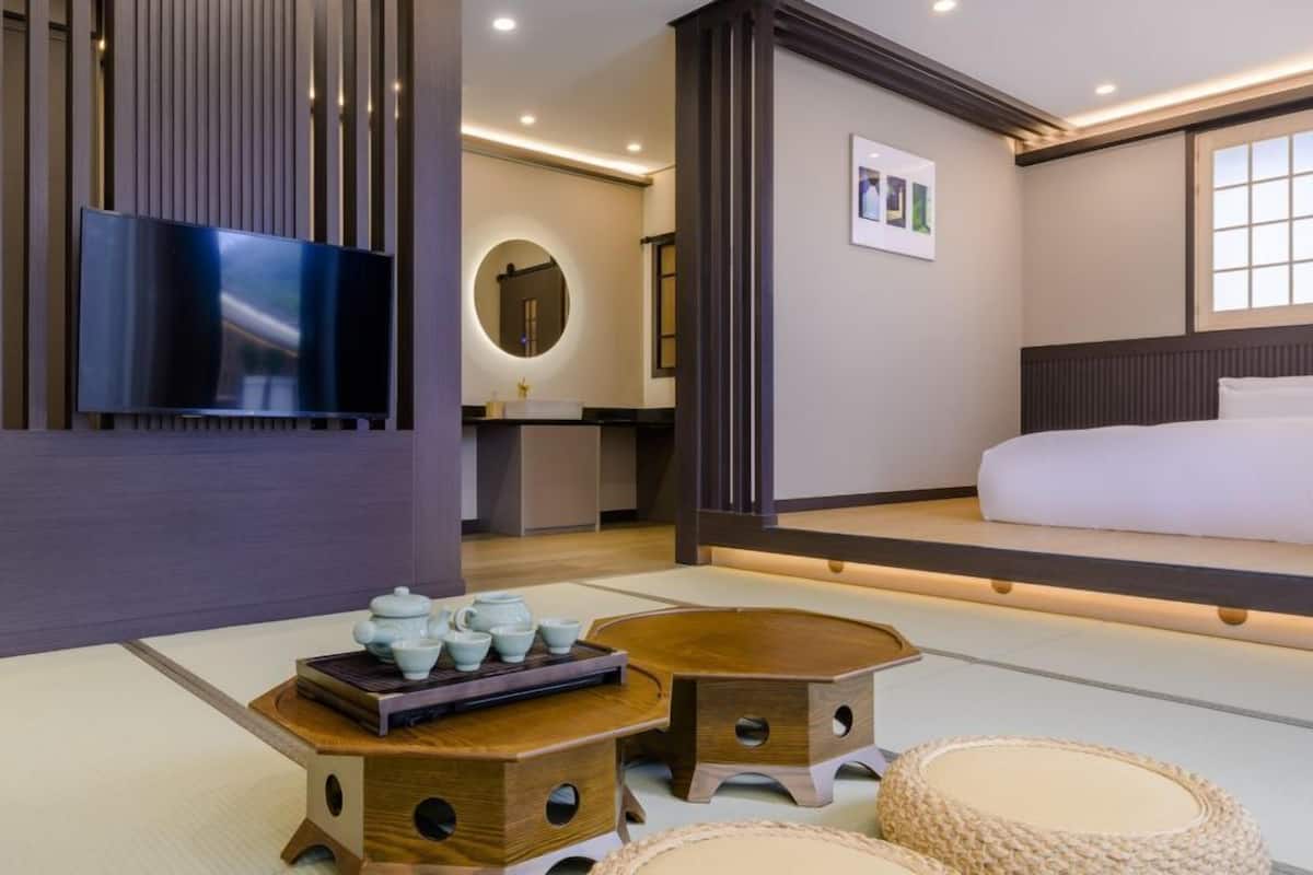 A modern hotel room with a bed, a TV, a small kitchenette, and a traditional Japanese tea set on the floor.