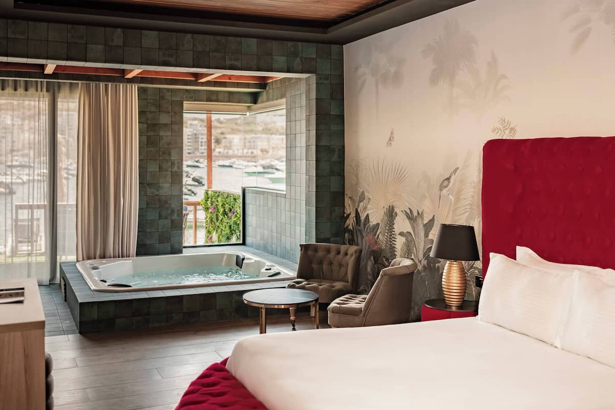 Quarto de hotel com uma cama grande, cabeceira vermelha, uma mesinha redonda, uma cadeira, mesa de cabeceira com abajur e uma banheira de hidromassagem.