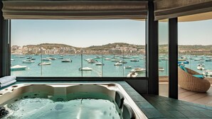 A luxurious room with a large window overlooking a marina filled with boats, a hot tub, and a wicker chair. - Damare Resort & Spa (St. Paul's Bay)