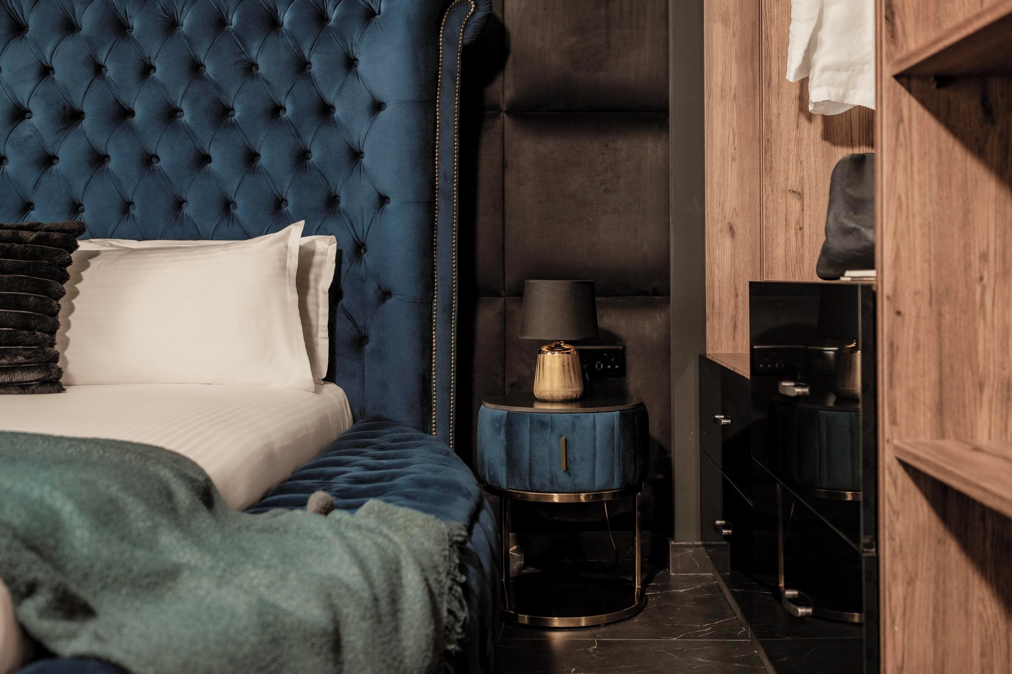 Een slaapkamer met een gestuufd hoofdeinde, een bed met witte lakens, een blauwe deken, een nachtkastje met een lamp en een houten garderobe.