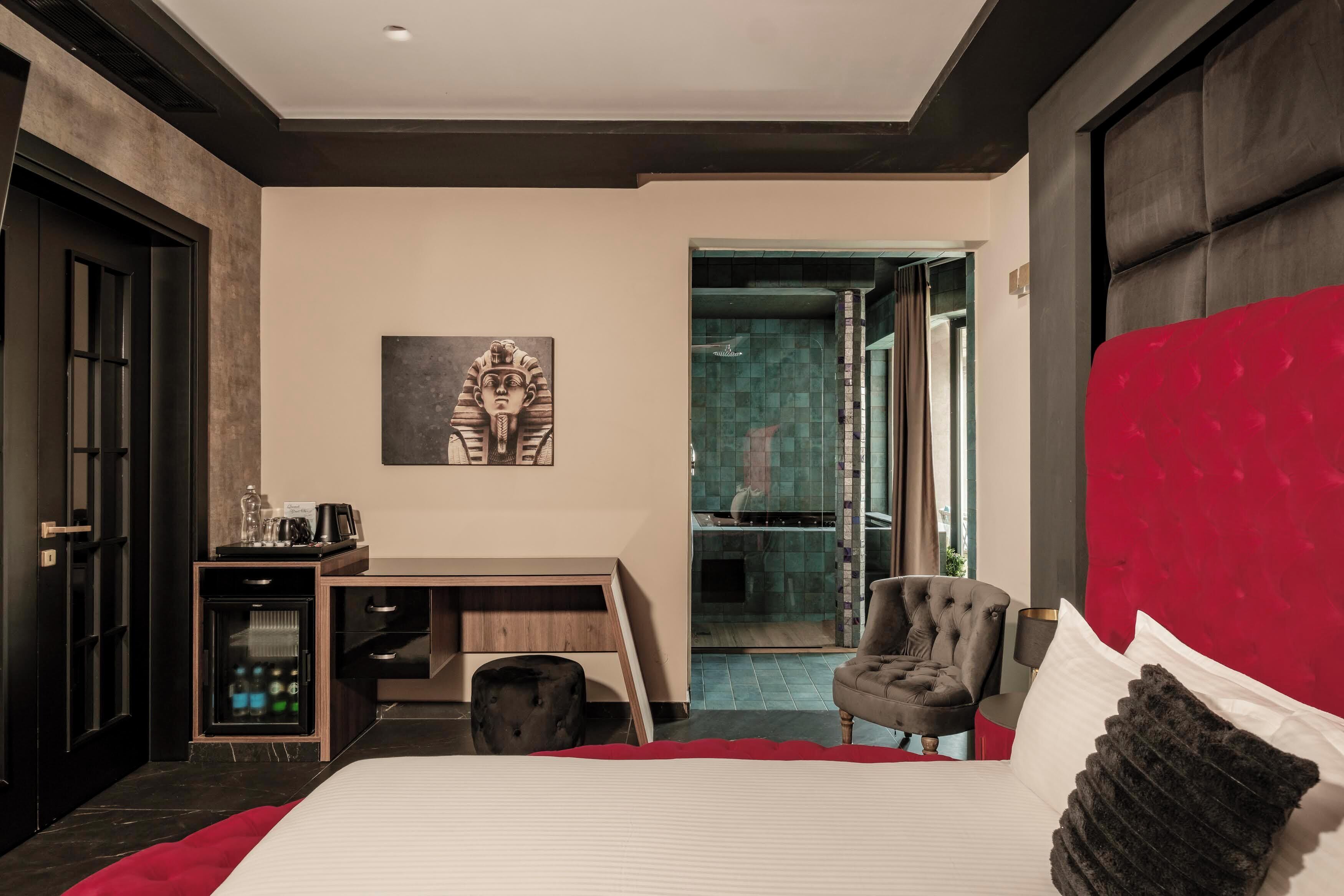 A modern hotel room with a large bed, a desk with a chair, a small refrigerator, and a red accent wall.