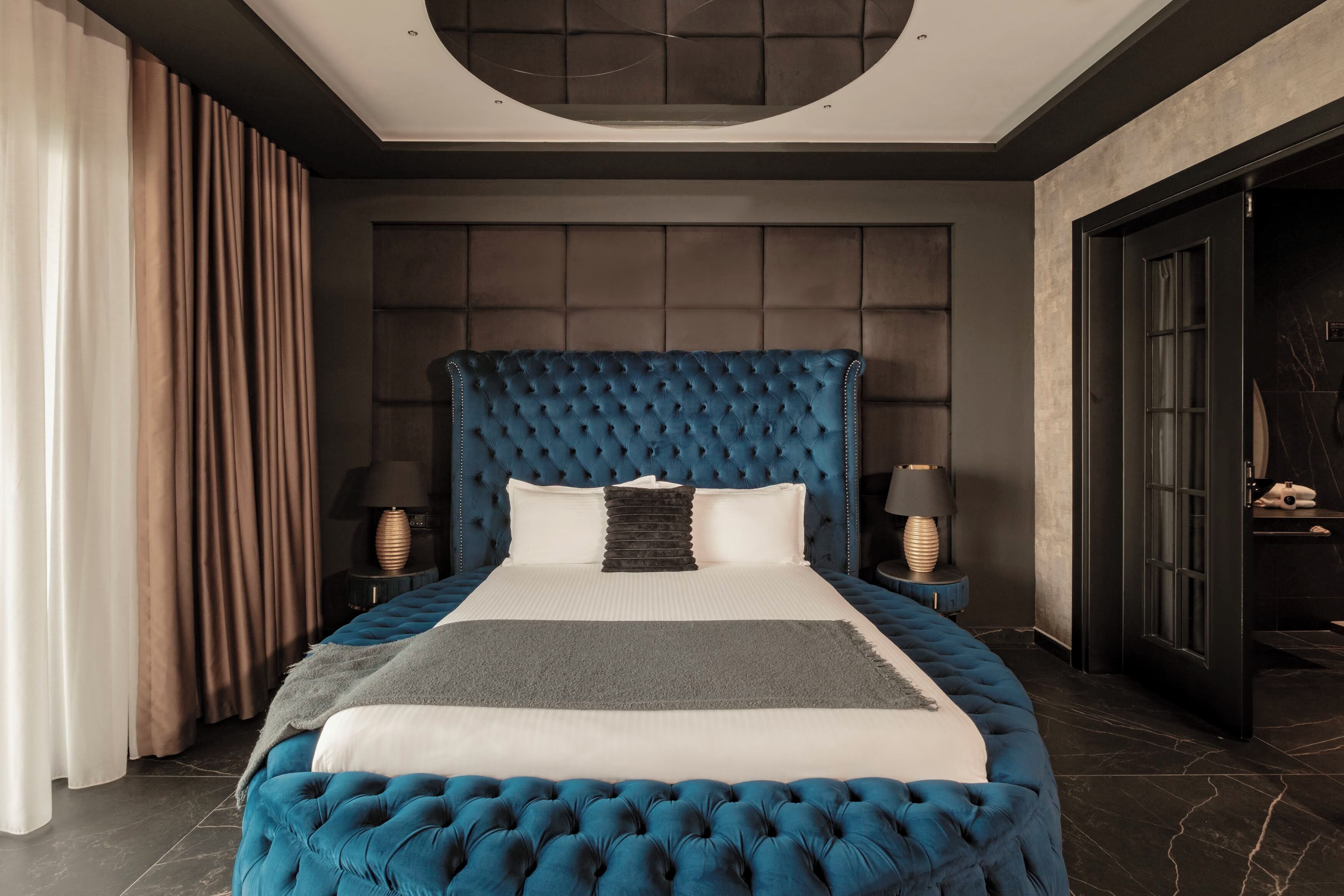 Uma cama grande e estofada em tom de azul, com roupa de cama branca e cinza, duas mesas de cabeceira com abajures, e um quarto com cores escuras e um detalhe circular no teto.