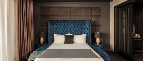 Uma cama grande e estofada em tom de azul, com roupa de cama branca e cinza, duas mesas de cabeceira com abajures, e um quarto com cores escuras e um detalhe circular no teto.