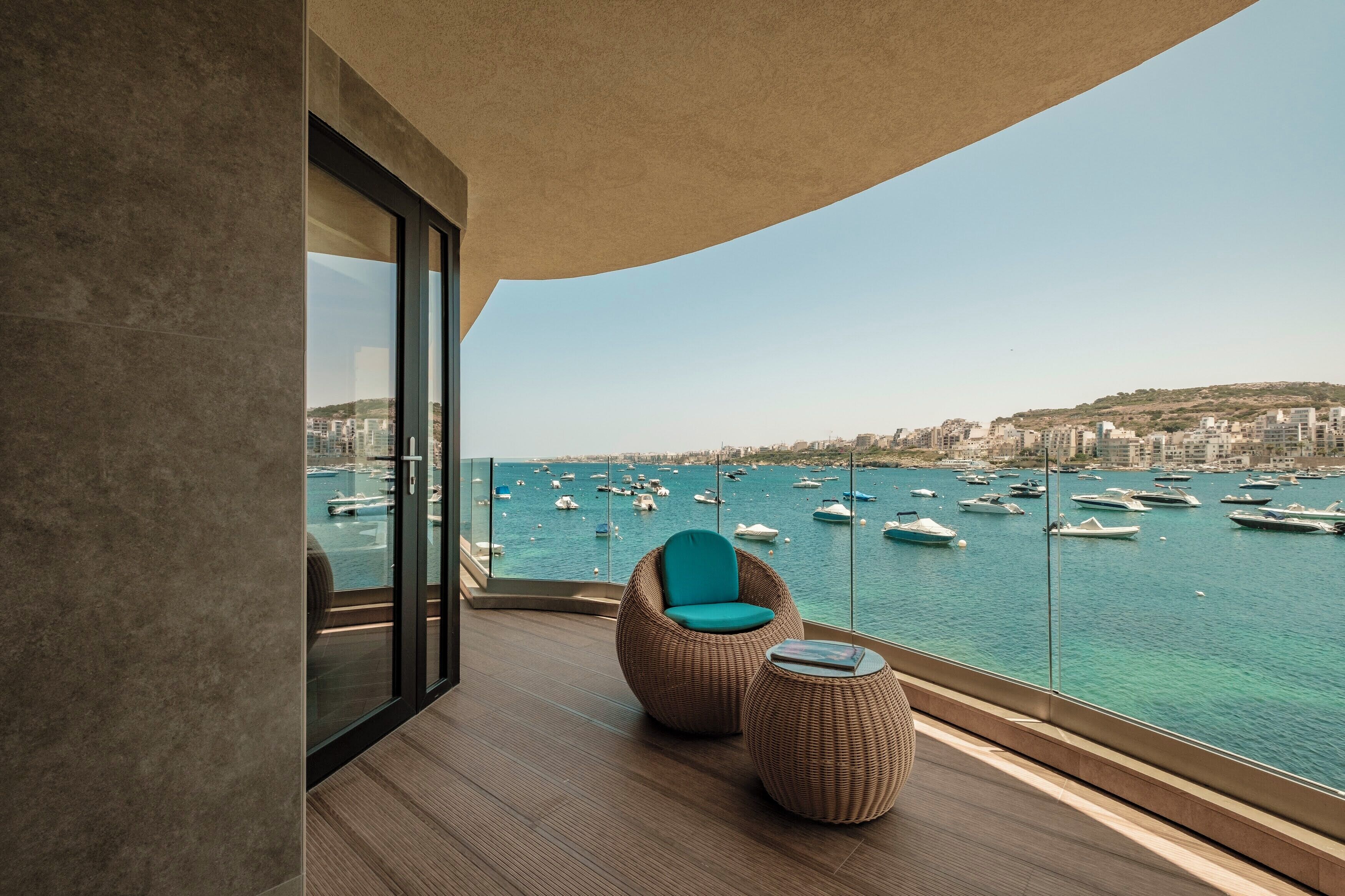 A balcony with a wicker chair and ottoman, overlooking a marina with boats and a cityscape.