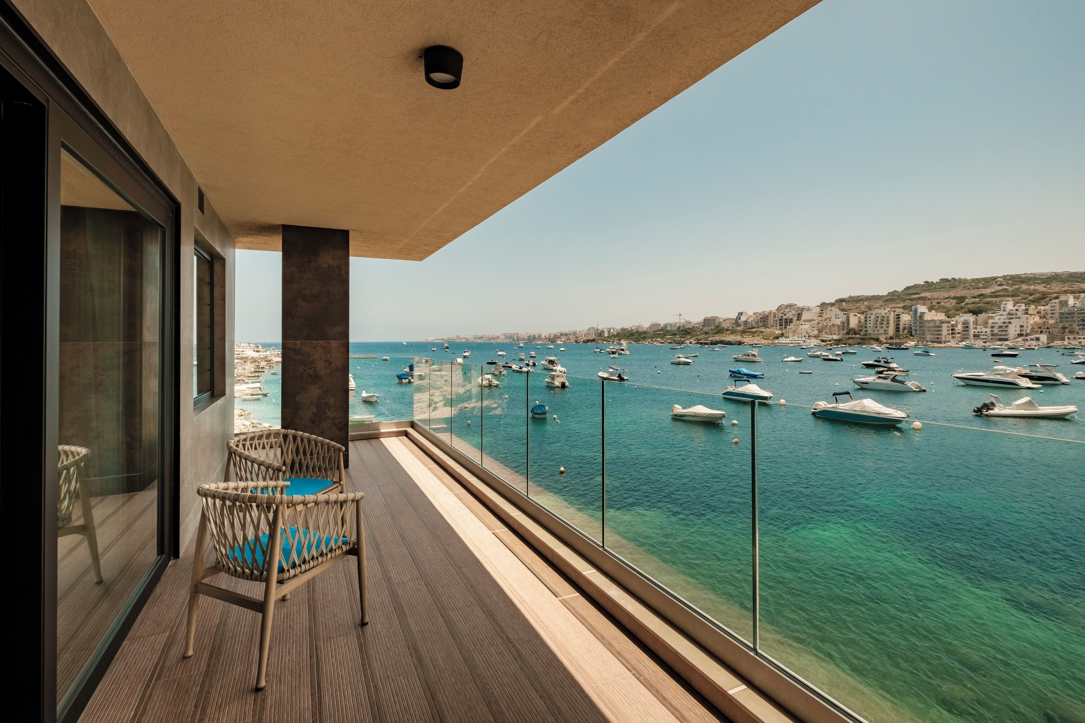 A balcony with a wicker chair, a view of a marina with boats, and a cityscape in the background.