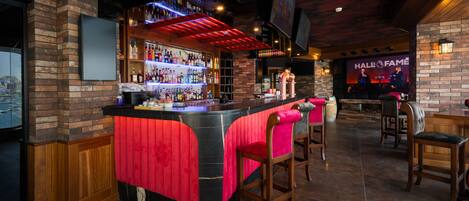 Una zona de bar con pared de ladrillo, una barra con taburetes y una pantalla que muestra 'Hall of Fame'.