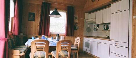 Une pièce confortable, aménagée comme un chalet, avec un intérieur en bois, une table à manger avec des chaises, un coin cuisine doté de meubles blancs et un luminaire suspendu.