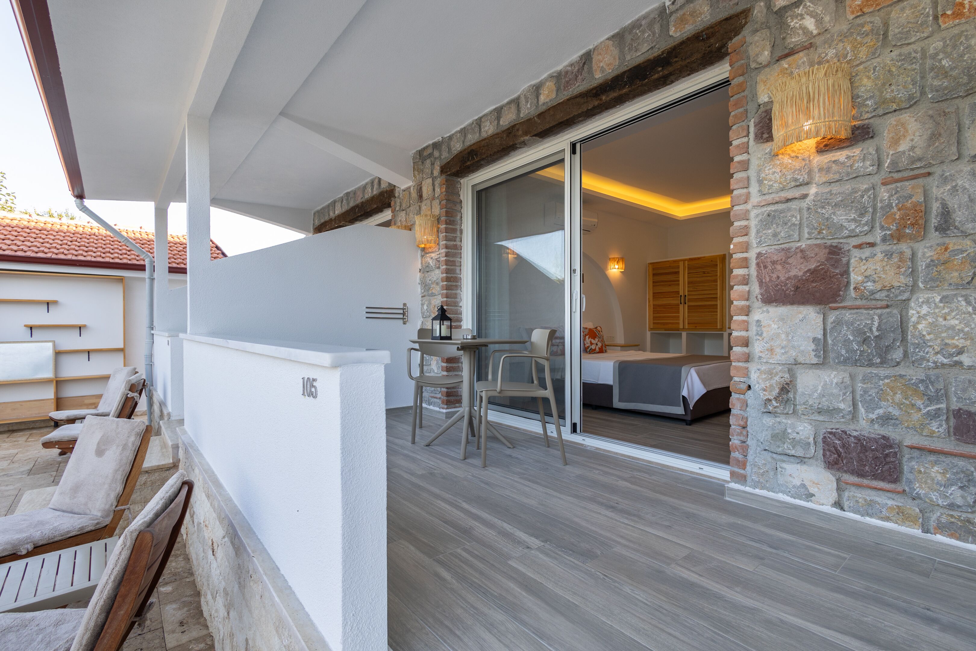 Un patio moderno al aire libre con un muro de piedra, un ventanal corredero que da a un dormitorio y una zona de estar con sillas y una mesa.