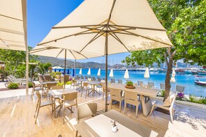 Un espacio al aire libre con mesas y sillas, grandes sombrillas y vistas a un puerto deportivo con barcos.