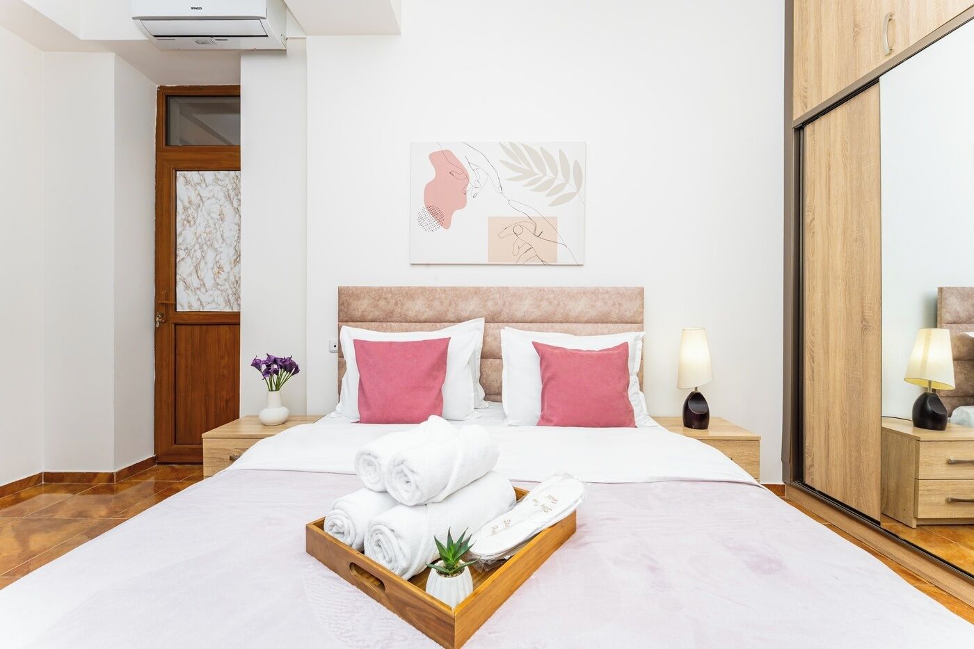 A hotel room with a bed, a wooden headboard, a nightstand with a lamp, a wooden door, and a wall-mounted artwork.