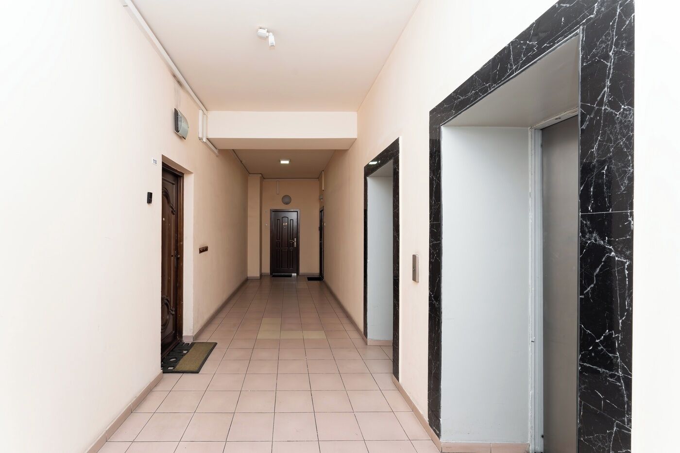 A hallway with a door at the end, a light fixture on the ceiling, and a tiled floor.