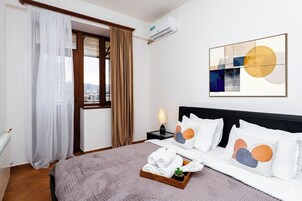 Een hotelkamer met een bed, een nachtkastje, een raam met gordijnen, een wandairconditioner en een lijst met een abstract schilderij.