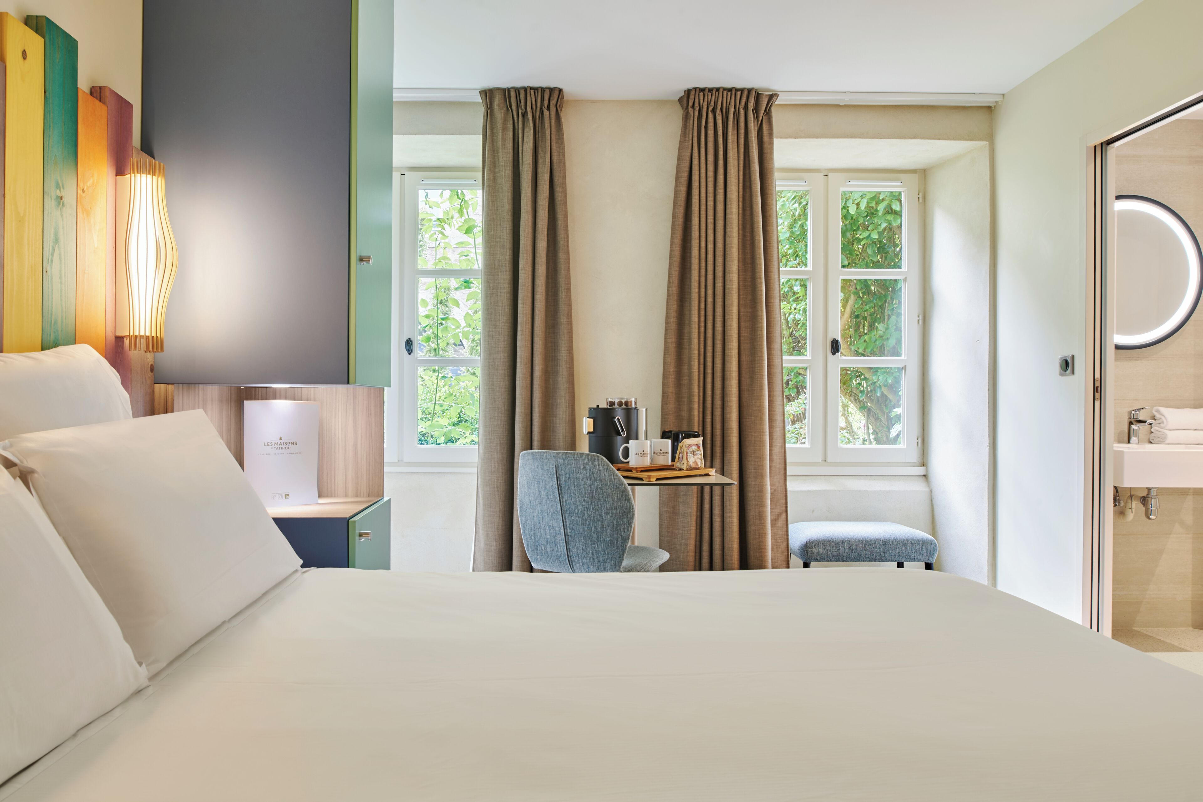 Une chambre d’hôtel comprenant un lit, un bureau, une chaise, une fenêtre avec des rideaux et une salle de bain avec un miroir.