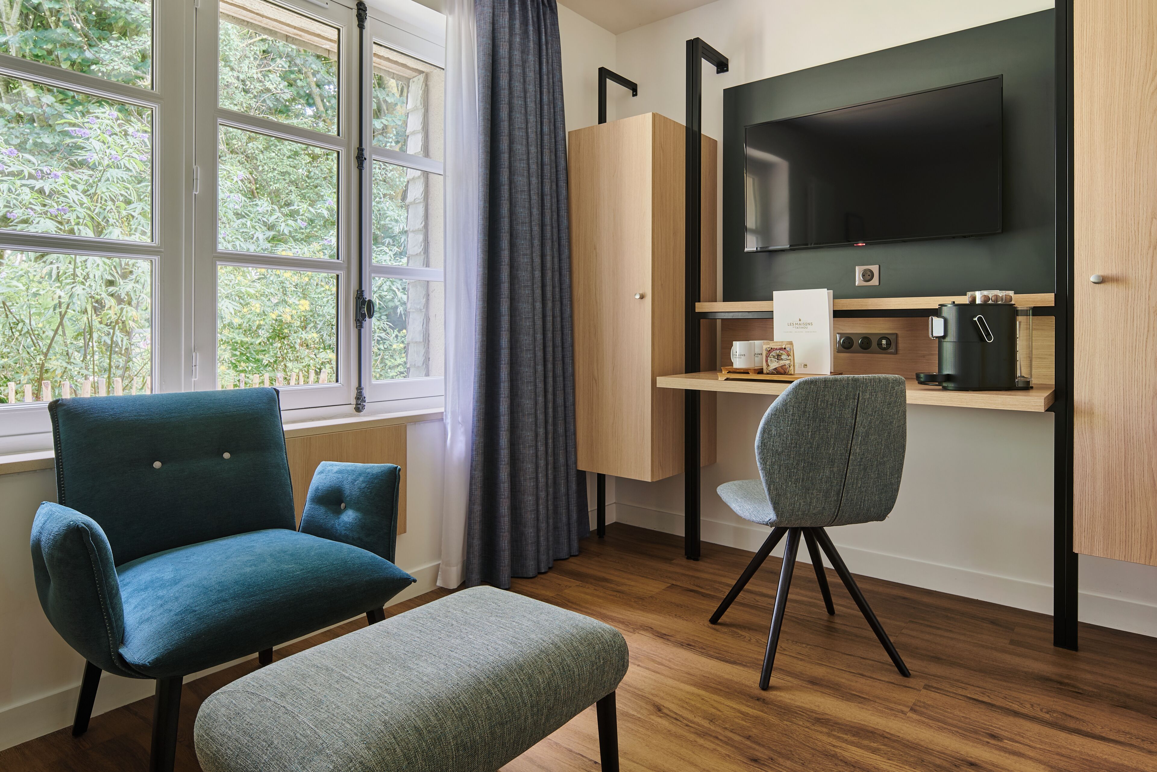 A room with a large window, a desk with a chair, a television, and a coffee machine.