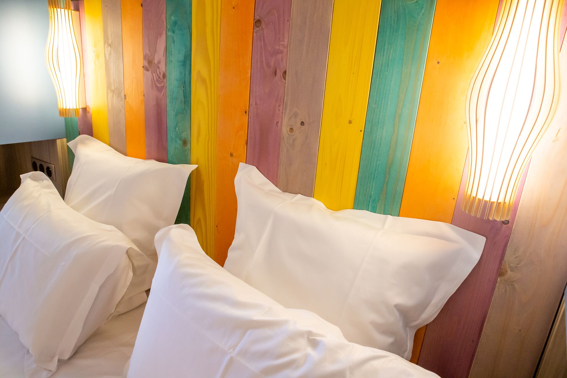 Un lit avec des oreillers blancs, adossé à un mur en bois aux couleurs variées.